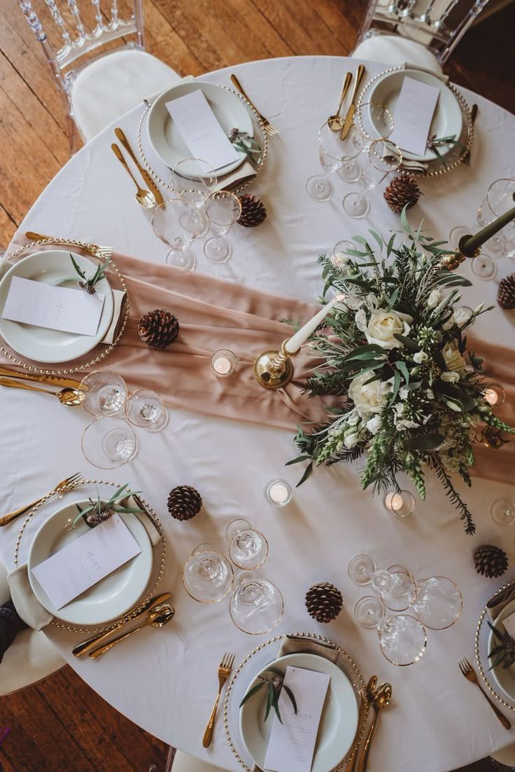 the table is set with white and gold place settings, silverware, pine cones, and greenery