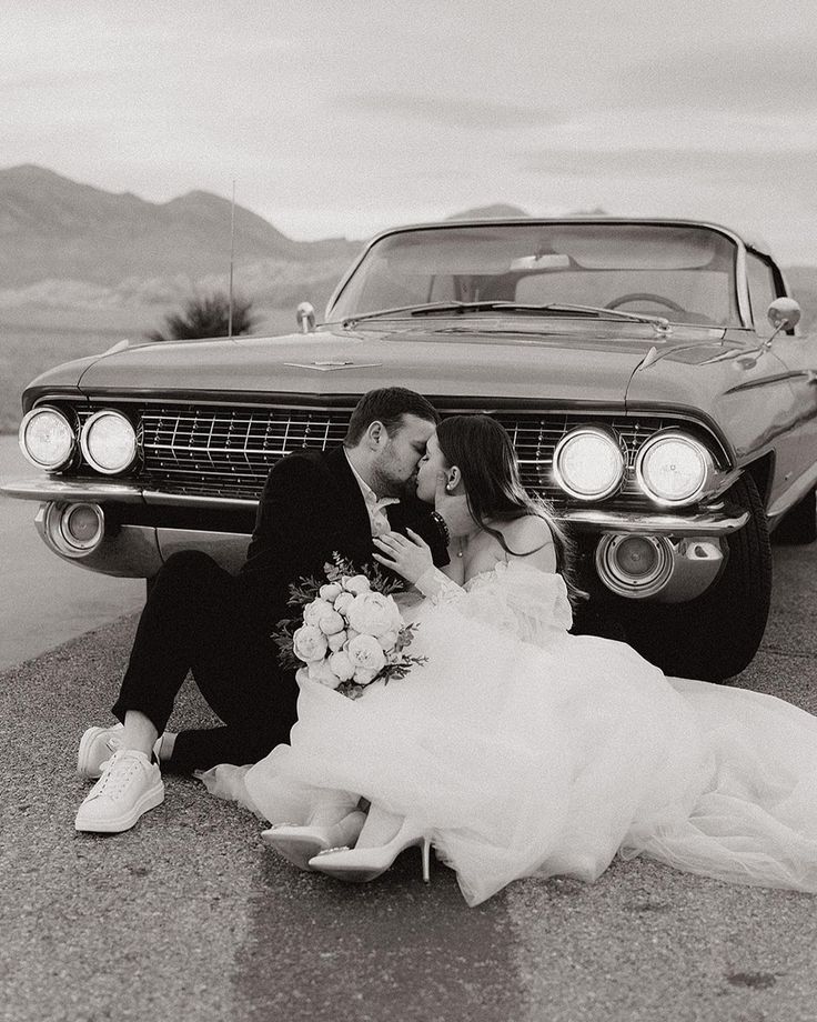 a bride and groom sitting on the ground next to a classic car in black and white