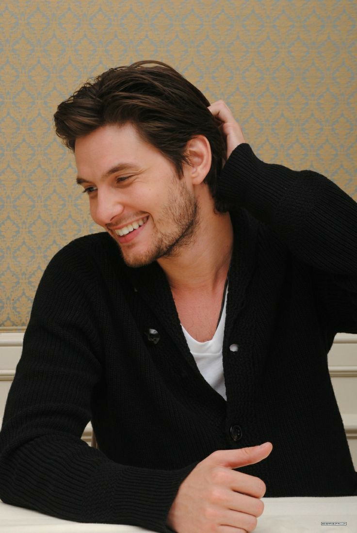 a man smiling while holding his hair in one hand and wearing a black cardigan