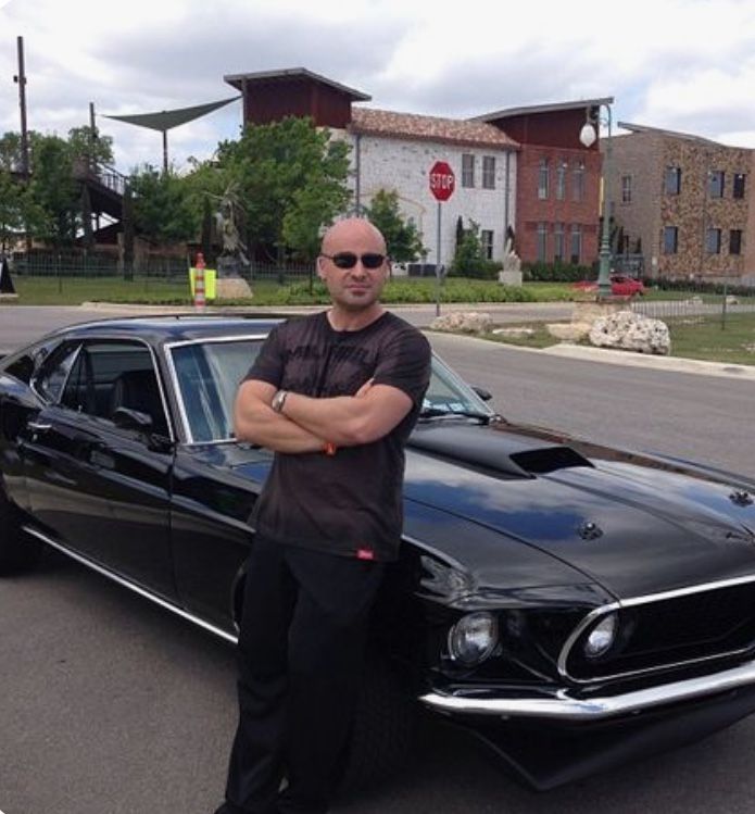 a man standing next to a black car in the middle of a parking lot with his arms crossed