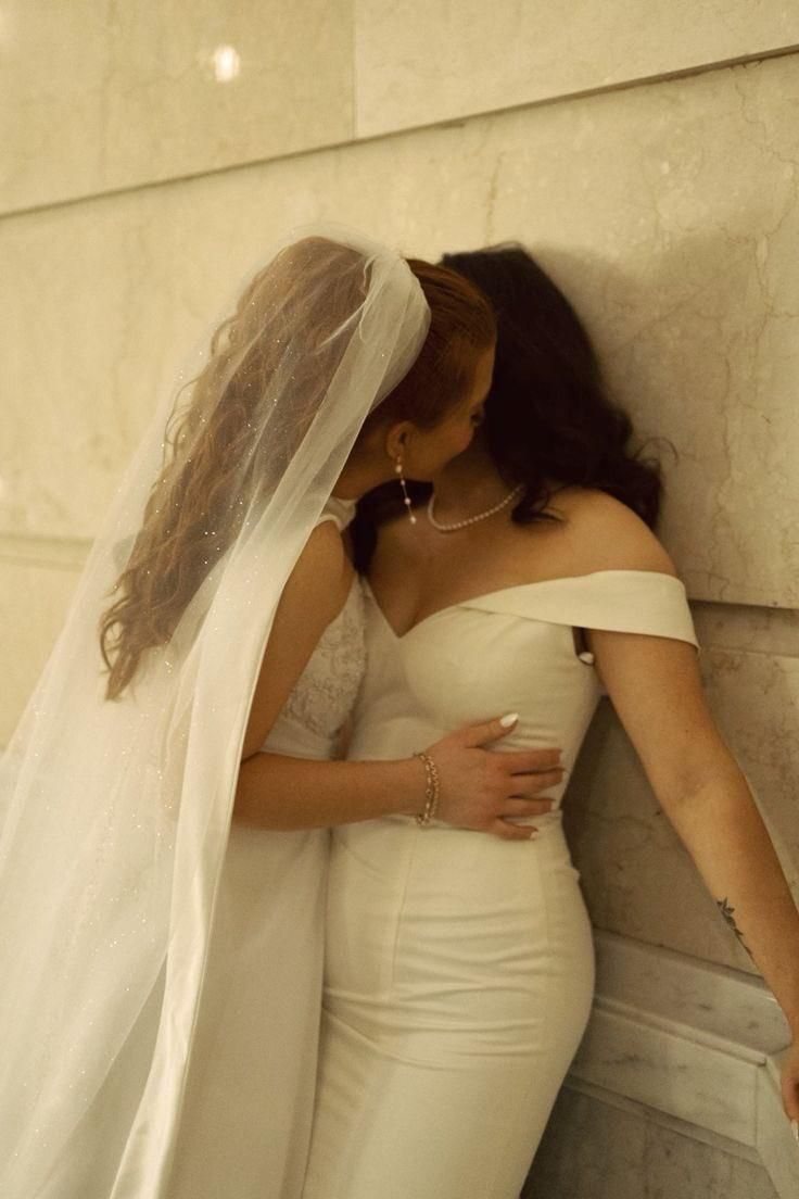 two women in white dresses and veils leaning against a wall with their hands on each other