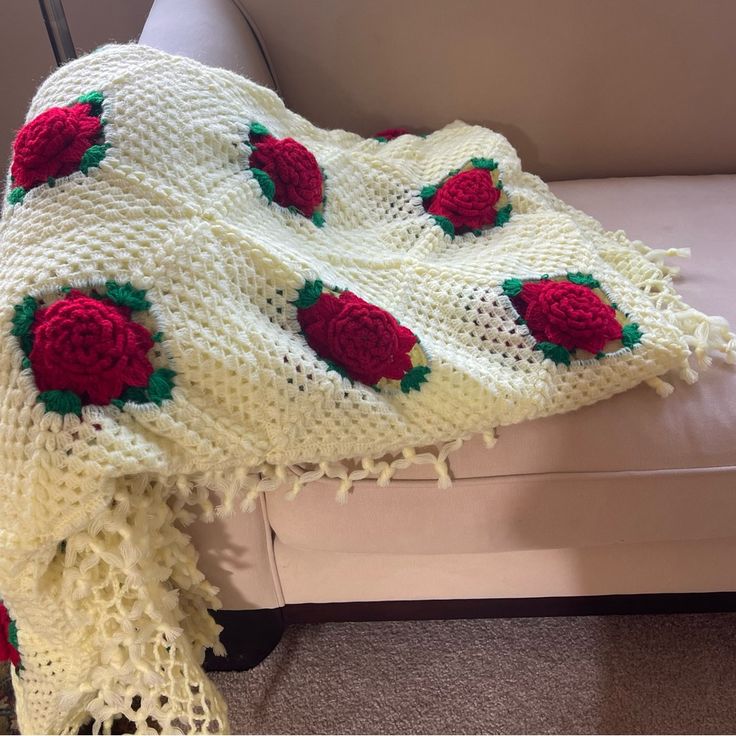 a white crocheted blanket with red roses on it sitting on a couch next to a pillow