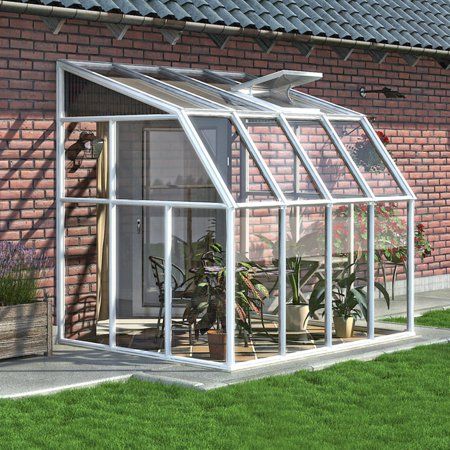 a small white greenhouse sitting on top of a green grass covered field next to a brick building