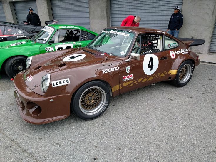 two racing cars parked next to each other in front of garage doors with people looking at them