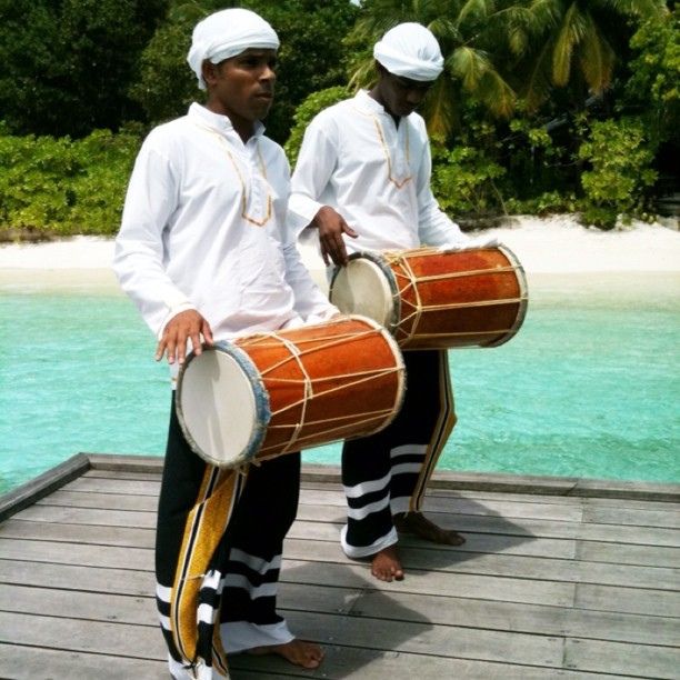 two men are playing drums on a dock near the water in front of some palm trees