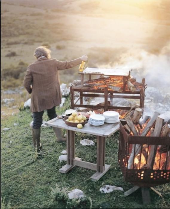 a woman cooking food over an open fire on top of a grass covered field with mountains in the background