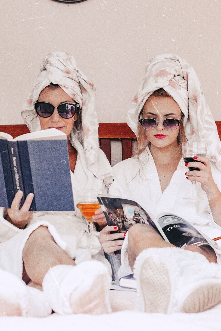 two women in bathrobes sitting on a bed reading books and drinking orange juice