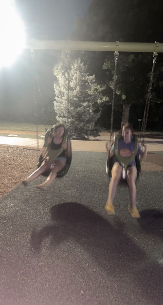 two people are swinging on swings in the park at night with bright lights behind them