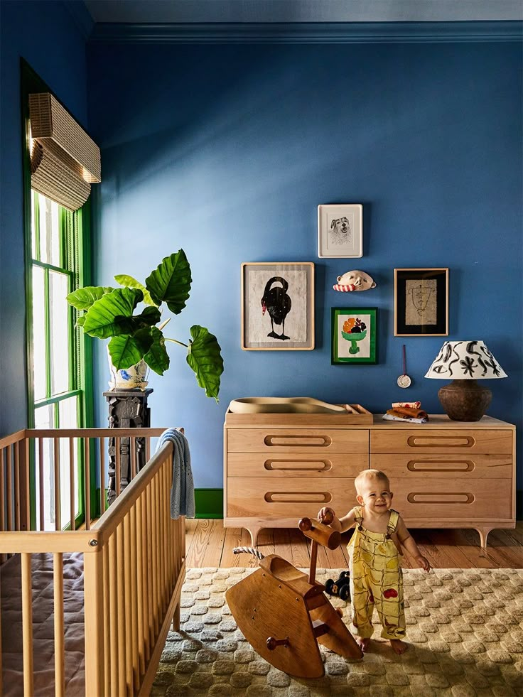 a baby standing next to a wooden rocking chair in a room with blue walls and pictures on the wall