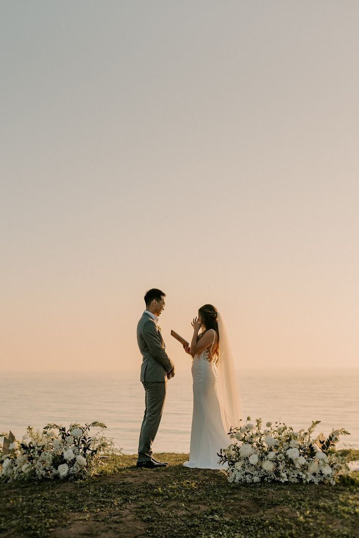 a bride and groom standing on top of a grass covered hill next to the ocean