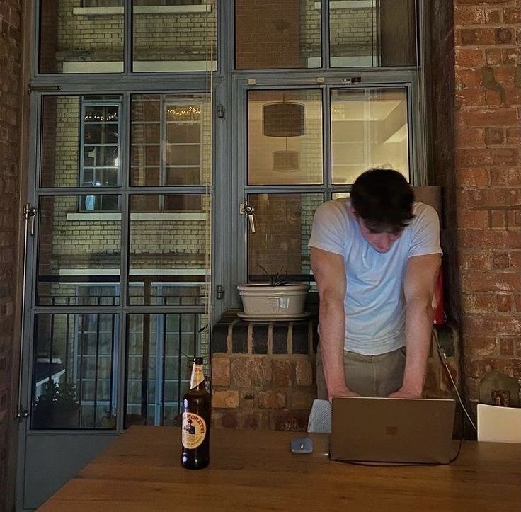 a man standing in front of a laptop computer on top of a wooden table next to a beer bottle