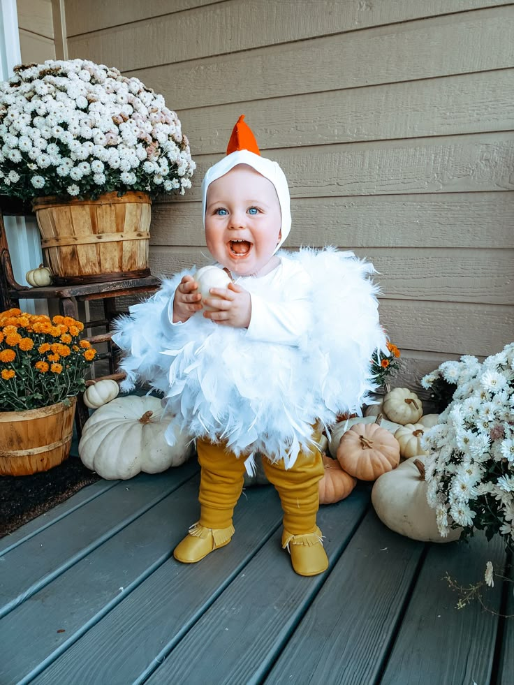 a baby dressed up as a chicken standing on a porch next to flowers and pumpkins