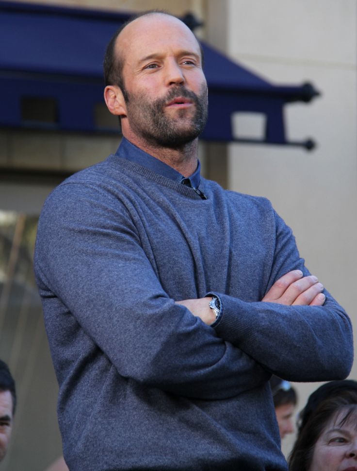 a man with his arms crossed standing in front of a building while people look on