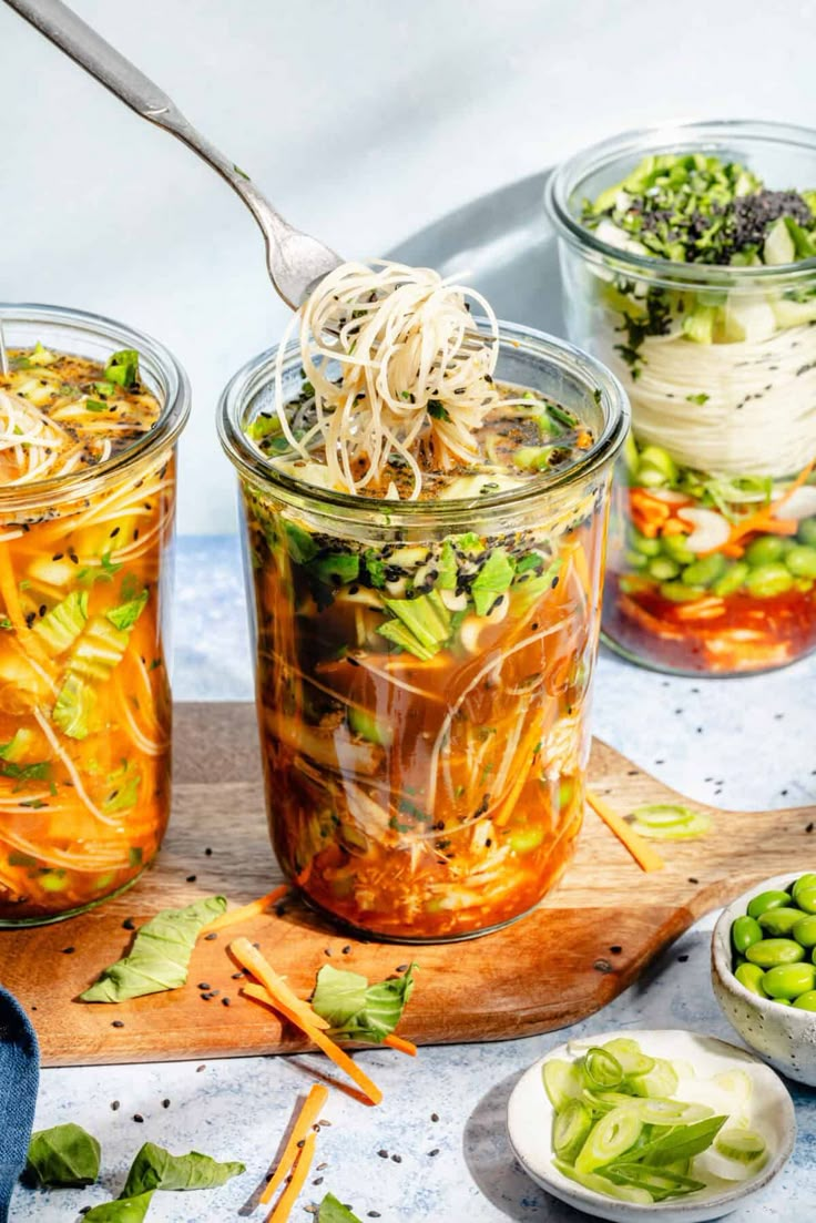 three jars filled with vegetables and noodles on top of a cutting board next to spoons