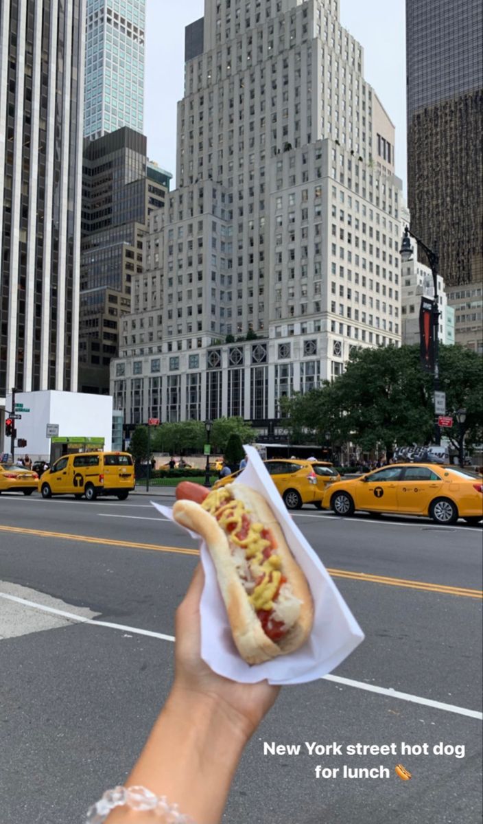 a person holding up a hot dog in the middle of a street with taxi cabs and tall buildings