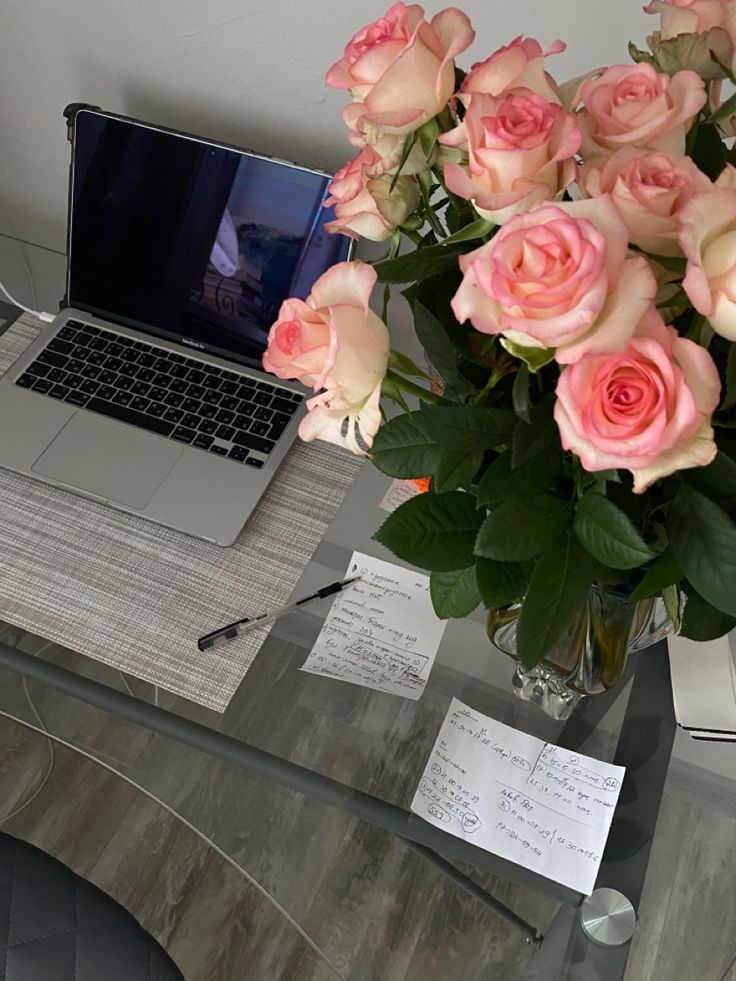 a vase filled with pink roses sitting on top of a desk next to a laptop computer