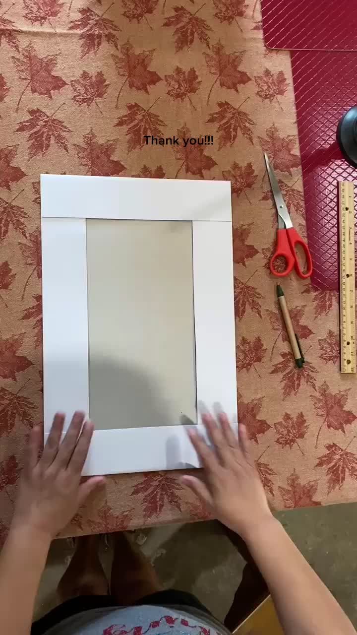 a person holding up a white frame with scissors and other items around it on a table