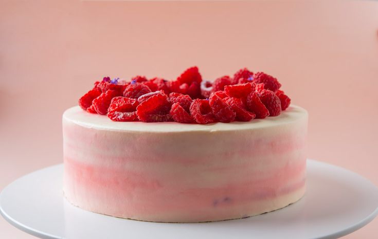 a cake with raspberries on top is sitting on a white plate against a pink background