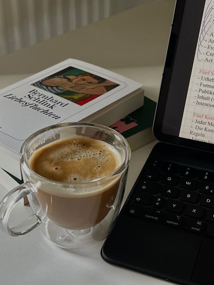 a cup of coffee next to a book and a laptop computer on a white table