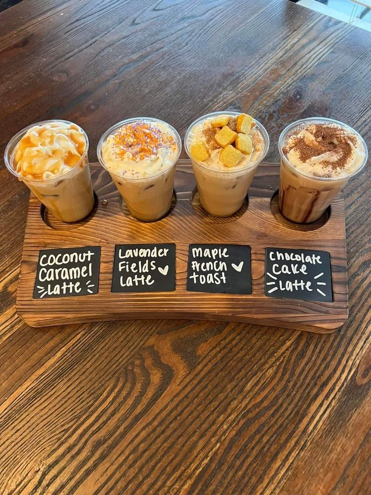 there are four different types of drinks on the wooden tray with chalkboard signs that read coconut caramel latte, macaroni salad and creme