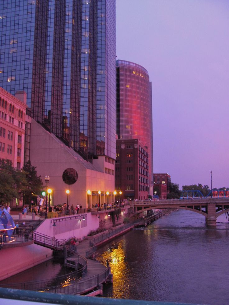 a river running through a city with tall buildings in the background at dusk, and people walking along it