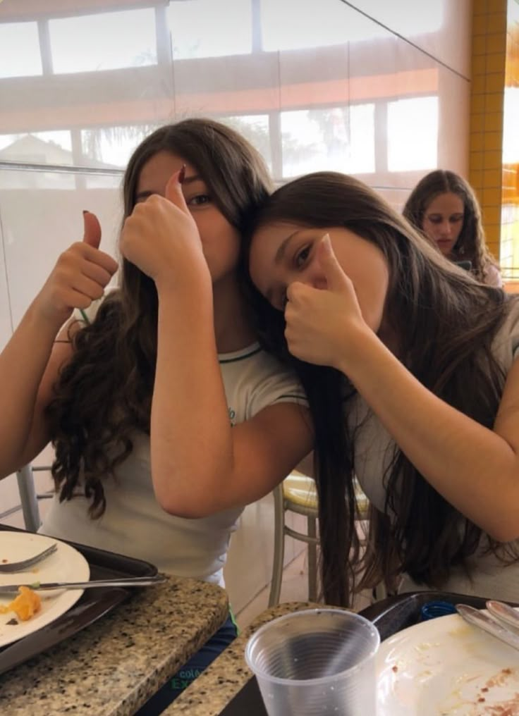 two young women sitting at a table with plates and forks in front of their mouths