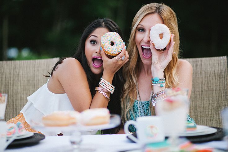 two women sitting at a table with doughnuts in front of their faces and one holding her mouth open