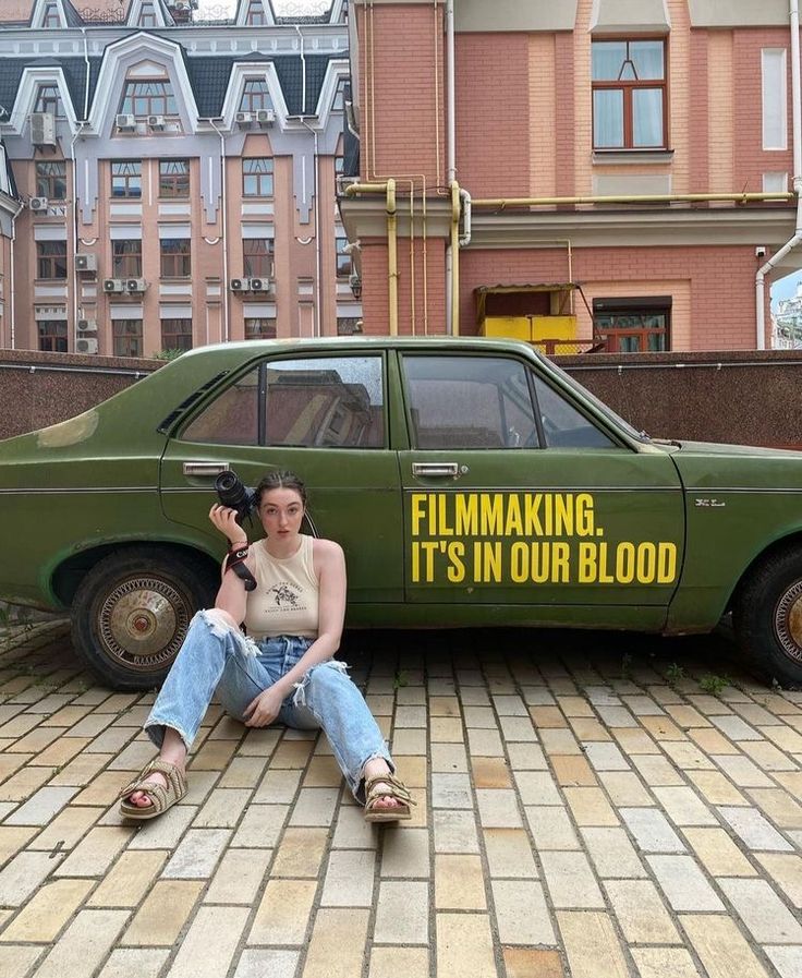 a woman sitting on the ground in front of a green car with an ad for film making it's in our blood