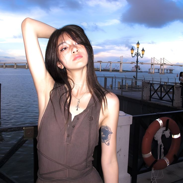 a woman in a brown dress standing next to the water with her arms behind her head