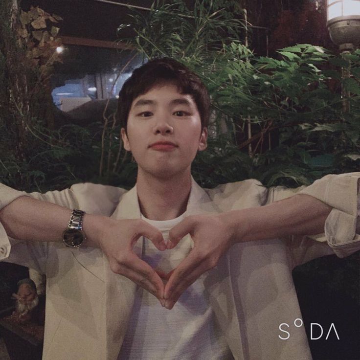 a man making a heart with his hands while standing in front of plants and greenery