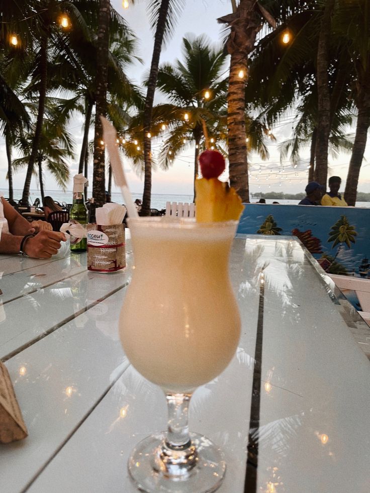 a drink sitting on top of a white table next to palm trees and the ocean