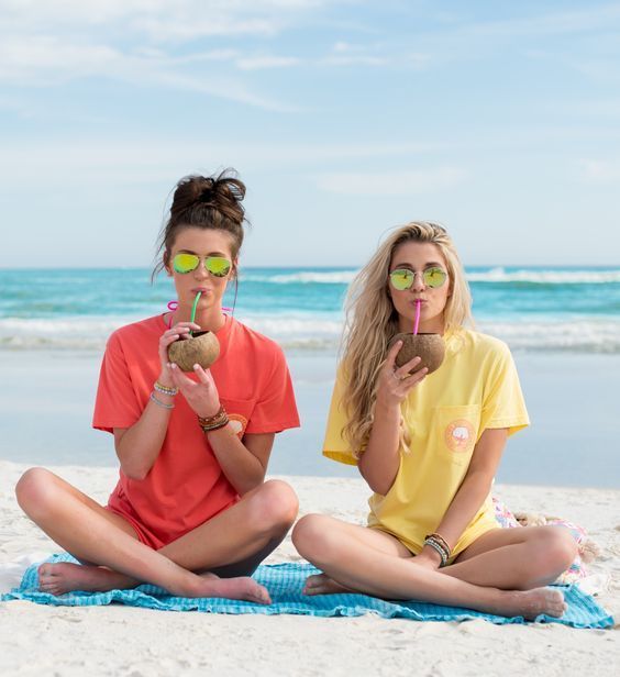 two young women sitting on the beach drinking beverages