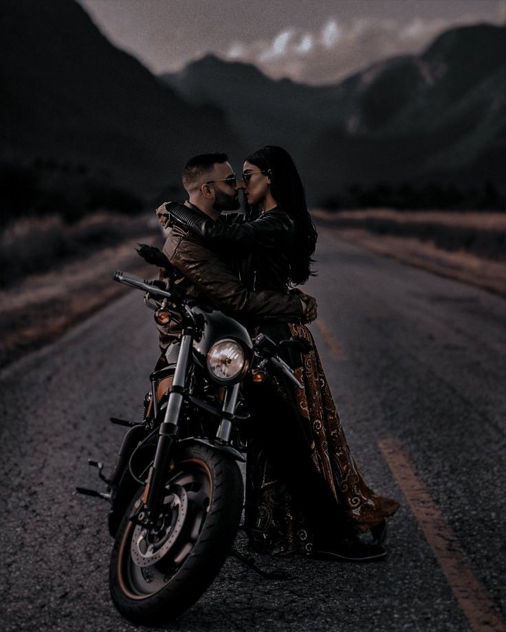 a man and woman sitting on a motorcycle in the middle of the road with mountains behind them