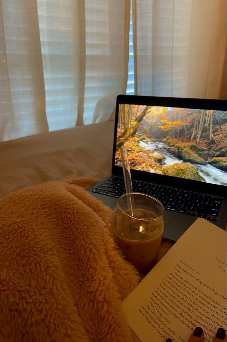 an open laptop computer sitting on top of a bed next to a book and drink