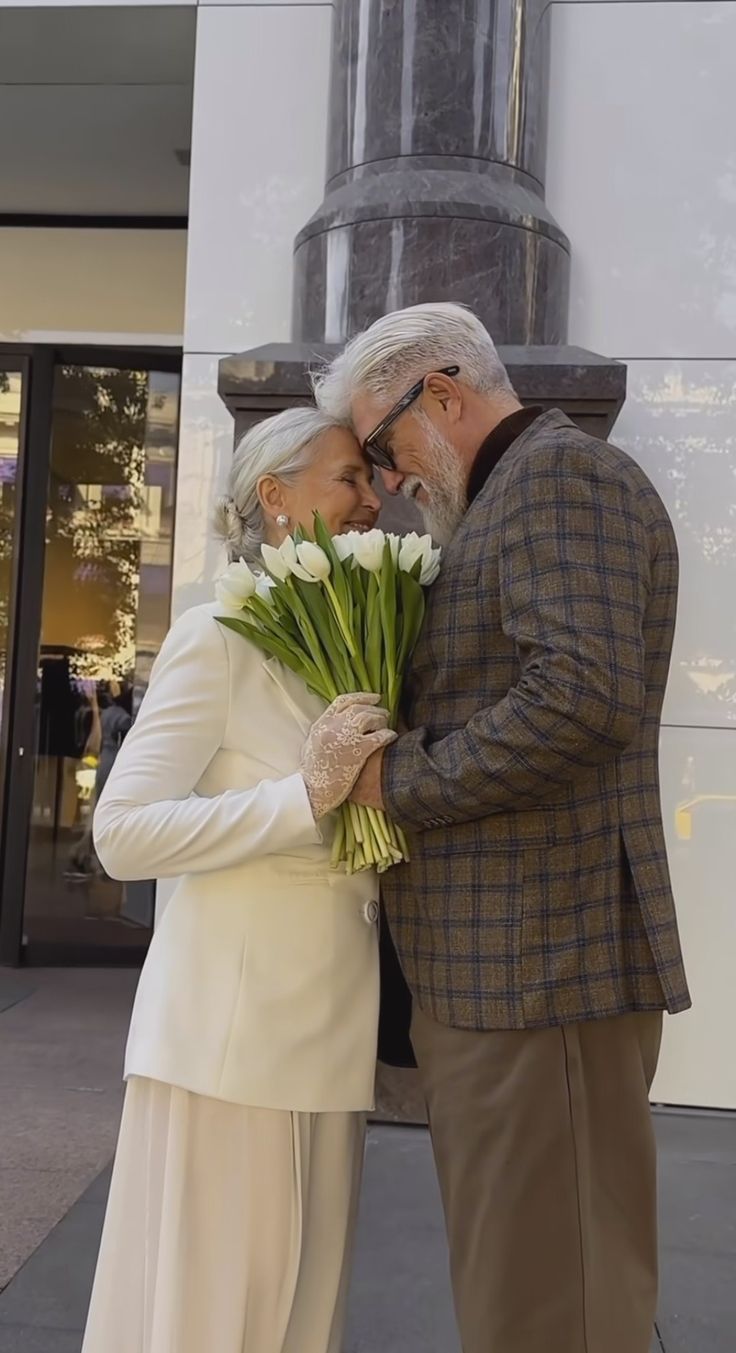 an older man and woman standing next to each other with flowers in front of them