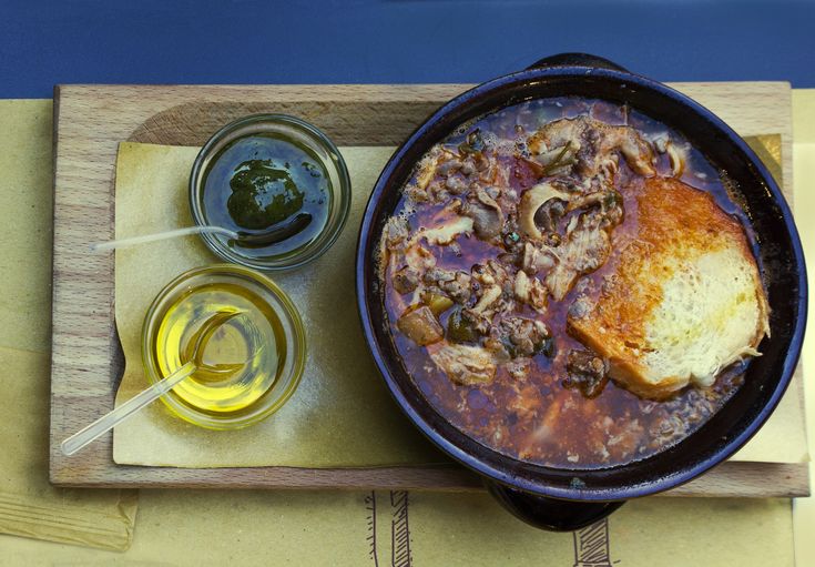 a bowl of stew with bread and olives on a cutting board next to some mustard