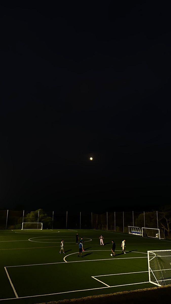 a group of people on a soccer field playing soccer at night with the moon in the sky