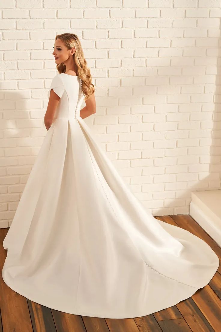 a woman wearing a white wedding dress standing in front of a brick wall with her back to the camera