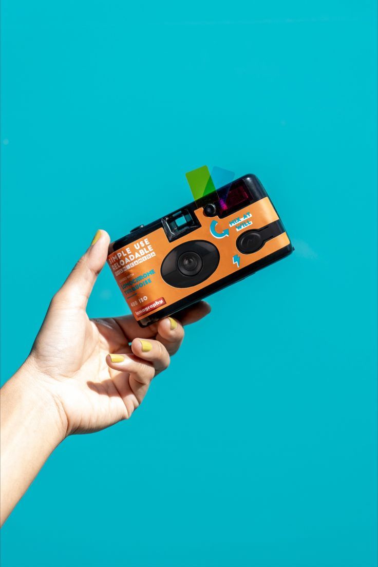 a hand holding an orange and black camera on top of a blue background with the lens up