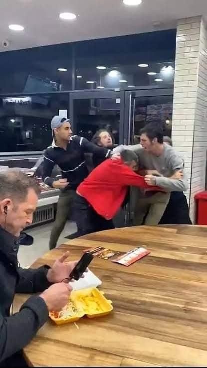 a man sitting at a table using his cell phone while other people stand in the background