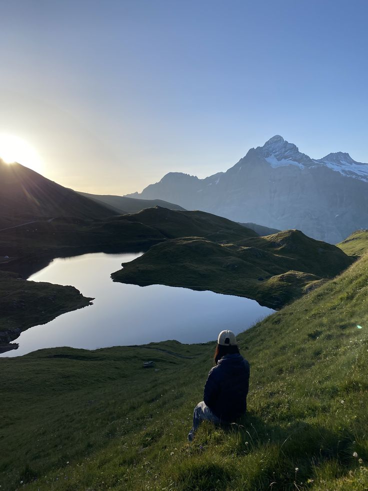 a man sitting on top of a grass covered hillside next to a lake in the mountains