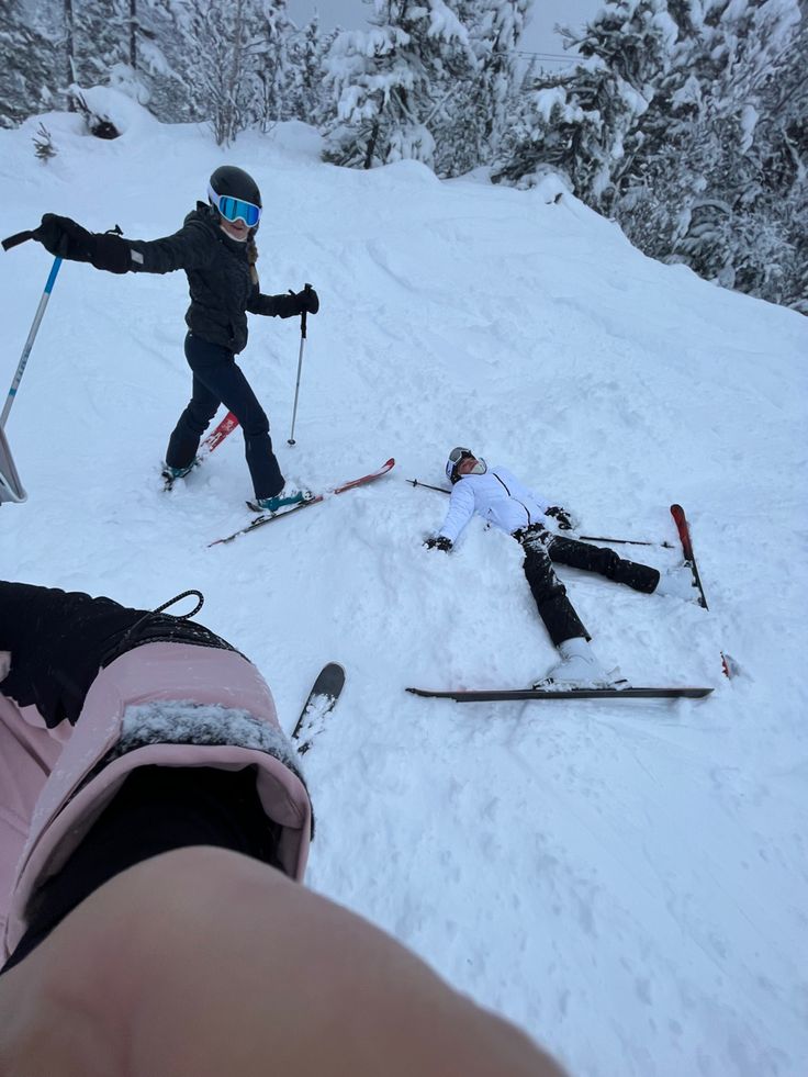 two people on skis in the snow with one laying down and another standing up