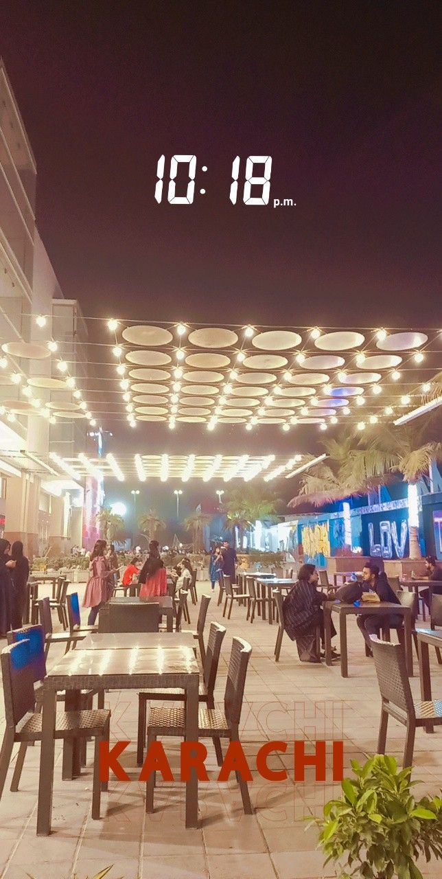 people are sitting at tables in the middle of an open air area with lights above them