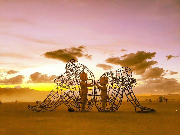 two people standing next to a metal sculpture in the middle of a field at sunset