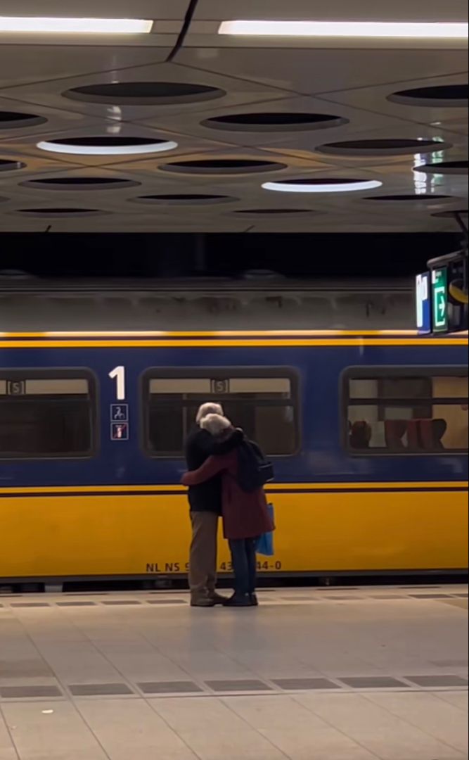 two people standing next to each other in front of a yellow and blue subway train