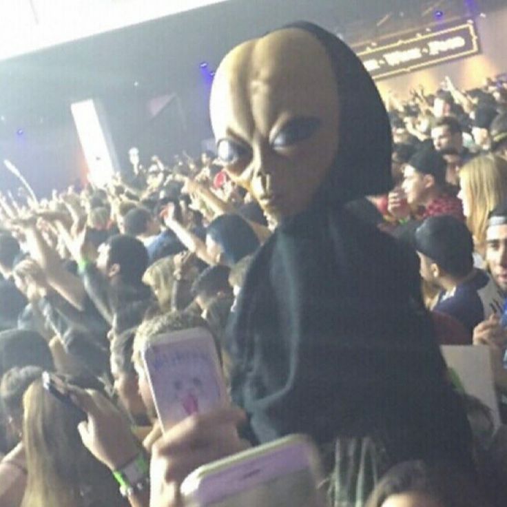 a person wearing an alien mask and holding a cell phone in front of a crowd