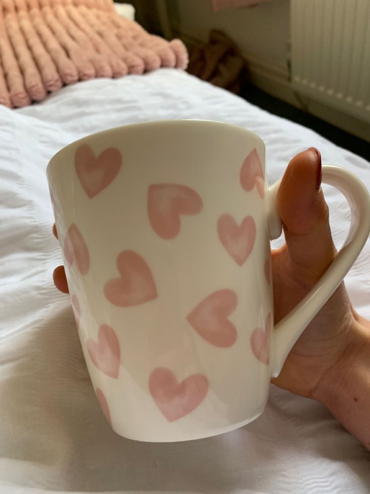 a hand holding a coffee cup with pink hearts on the outside and inside, in front of a white bed