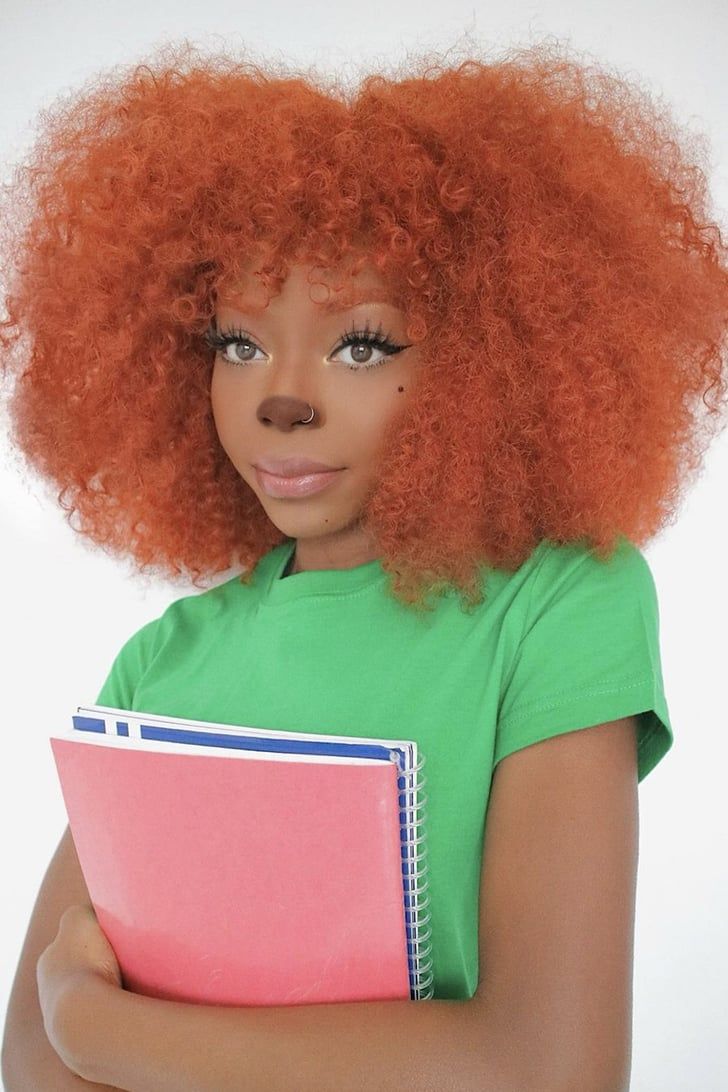 a woman with an orange afro holding a pink binder in her hand and looking at the camera