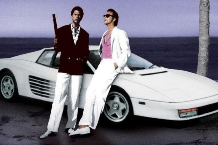 two men in suits standing next to a white sports car on the beach with palm trees