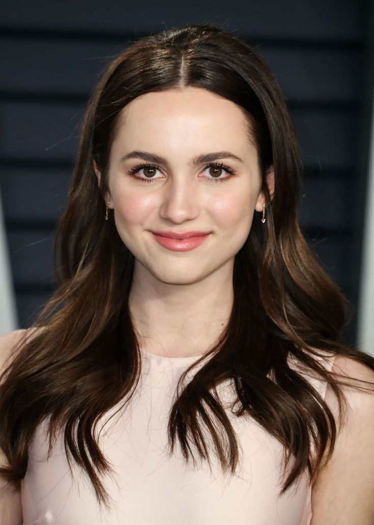 a woman with long brown hair wearing a white dress and smiling at the camera while standing in front of a wall